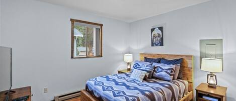 Un dormitorio con una cama de madera, dos mesitas de noche con lámparas, una ventana y un cuadro en la pared.