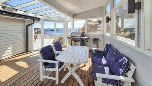 A covered outdoor patio with a table and chairs, a grill, and a view of the sea.