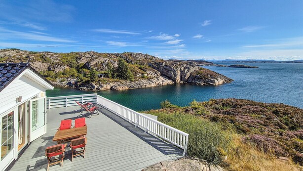 A coastal property with a wooden deck, outdoor seating, and a view of the sea and rocky landscape.