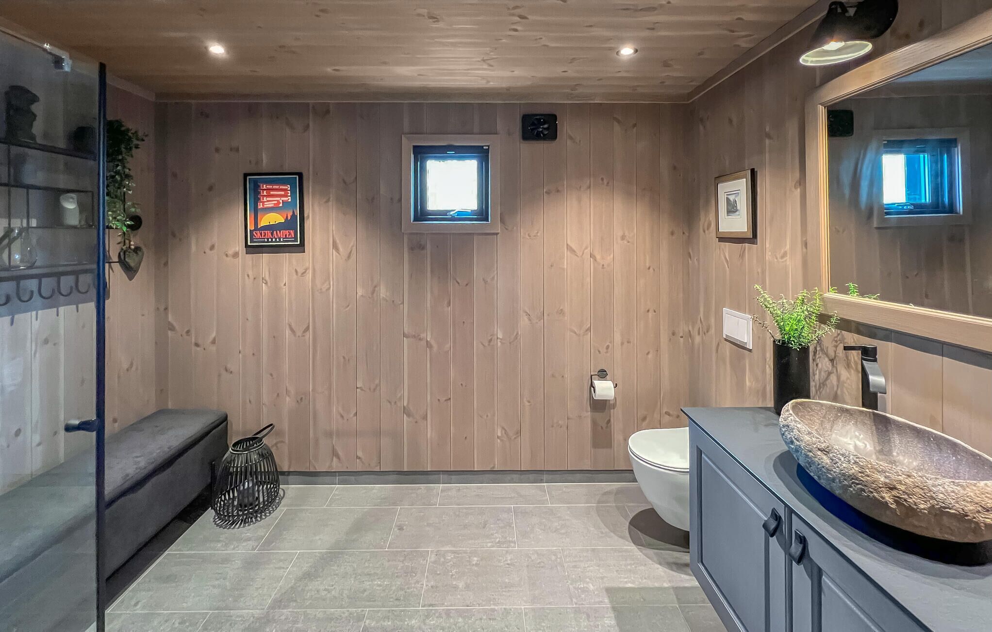 Une salle de bain aux murs lambrissés, équipée de toilettes, d’un lavabo et d’un miroir. Un tableau encadré est accroché au mur, et une petite fenêtre se trouve au-dessus des toilettes.