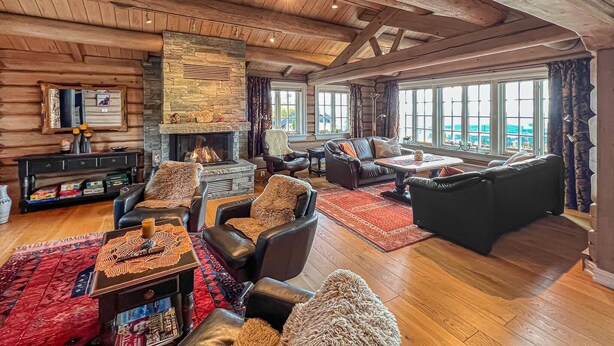 A cozy log cabin interior with a stone fireplace, leather furniture, and large windows offering a view of the outdoors.