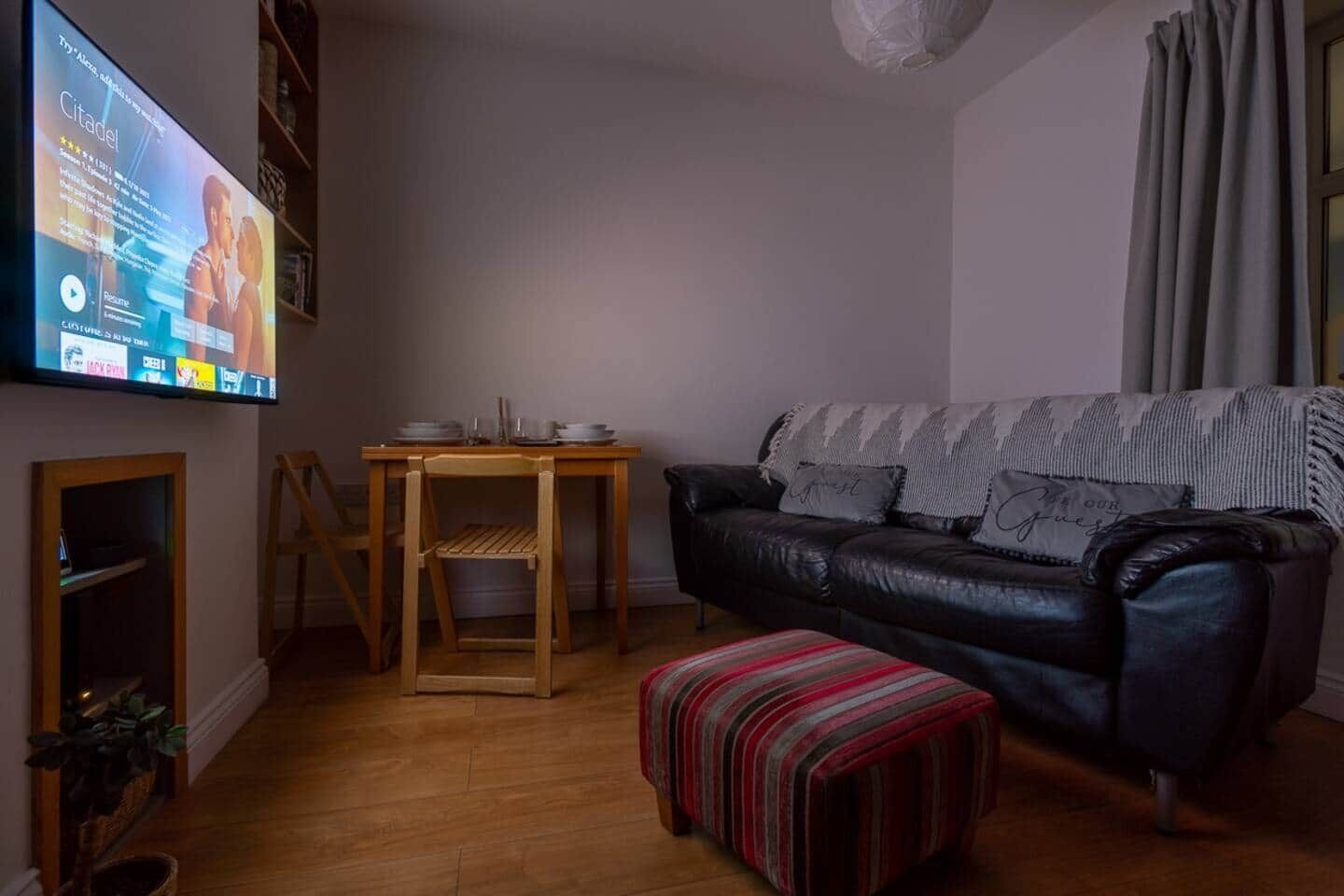 A living room with a black leather sofa, a wooden table, and a television mounted on the wall.