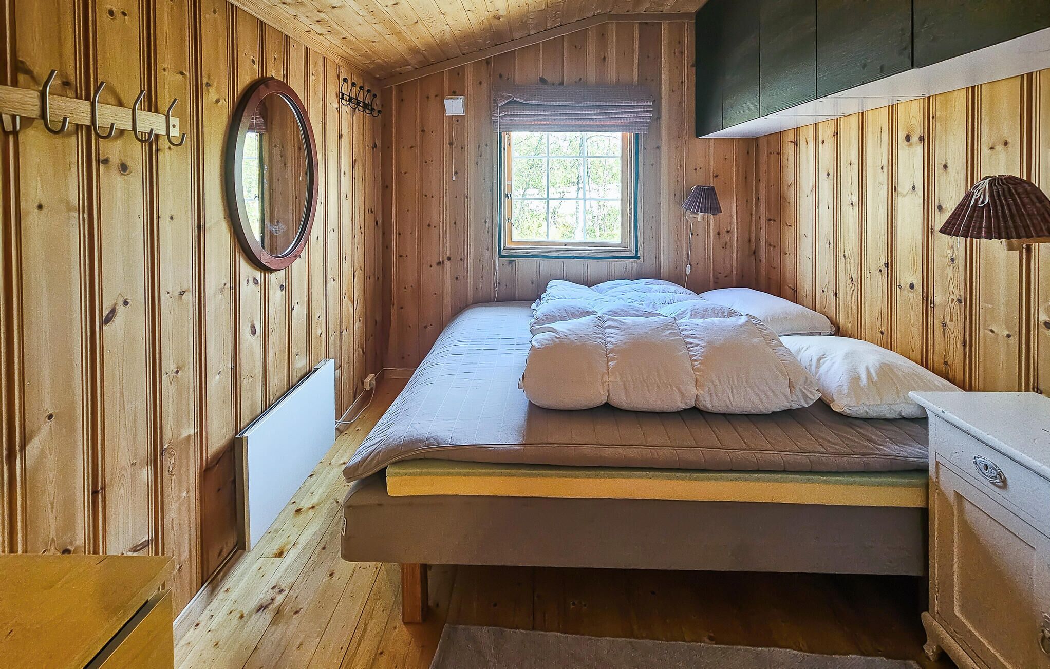 A wooden cabin bedroom with a single bed, a window with a view, and a wooden dresser.