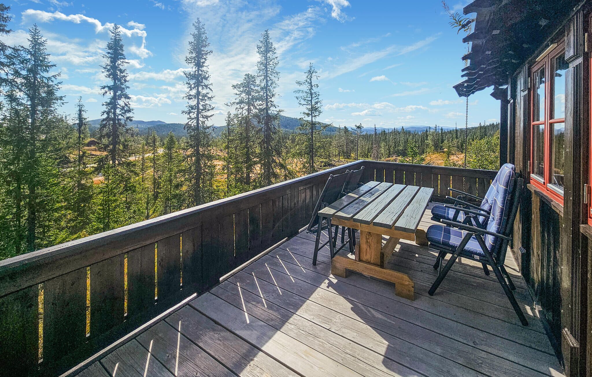 Eine Holzdielen-Terrasse mit Tisch und Stühlen, mit Blick auf eine bewaldete Landschaft und Berge in der Ferne.