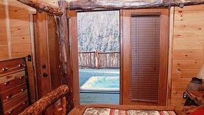 A wooden cabin room with a bed, a wooden bench, and a view of a hot tub outside. - 3-Bedroom Master Deluxe (Chapmanville)