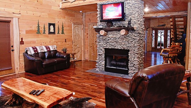 A log cabin interior with a stone fireplace, a leather sofa, a wooden coffee table, and a television mounted above the fireplace. - 3-Bedroom Master Deluxe (Chapmanville)
