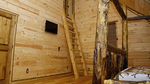 Ein Holzblockhaus-Interieur mit einer Holzleiter, einem an der Wand montierten Fernseher und einem Bett.