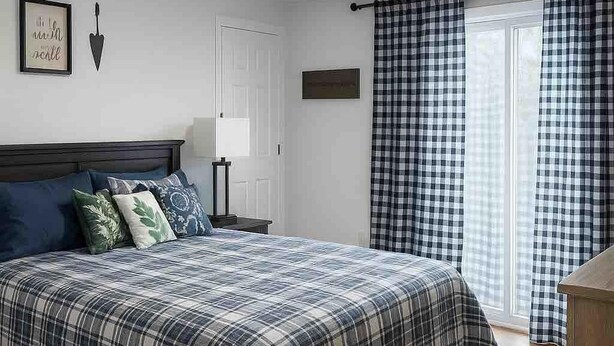 A bedroom with a bed, a nightstand, a lamp, a door, and a window with checkered curtains. - Creek Side Retreat (Chapmanville)