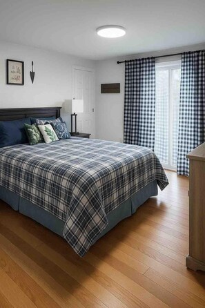 A bedroom with a bed, a nightstand, a lamp, a door, and a window with checkered curtains. - Creek Side Retreat (Chapmanville)