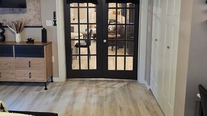 Ein Zimmer mit Holzboden, einem Bett mit weißer Bettdecke, einer schwarzen Tür mit Fenster, einem an der Wand montierten Fernseher und einem Deckenventilator.