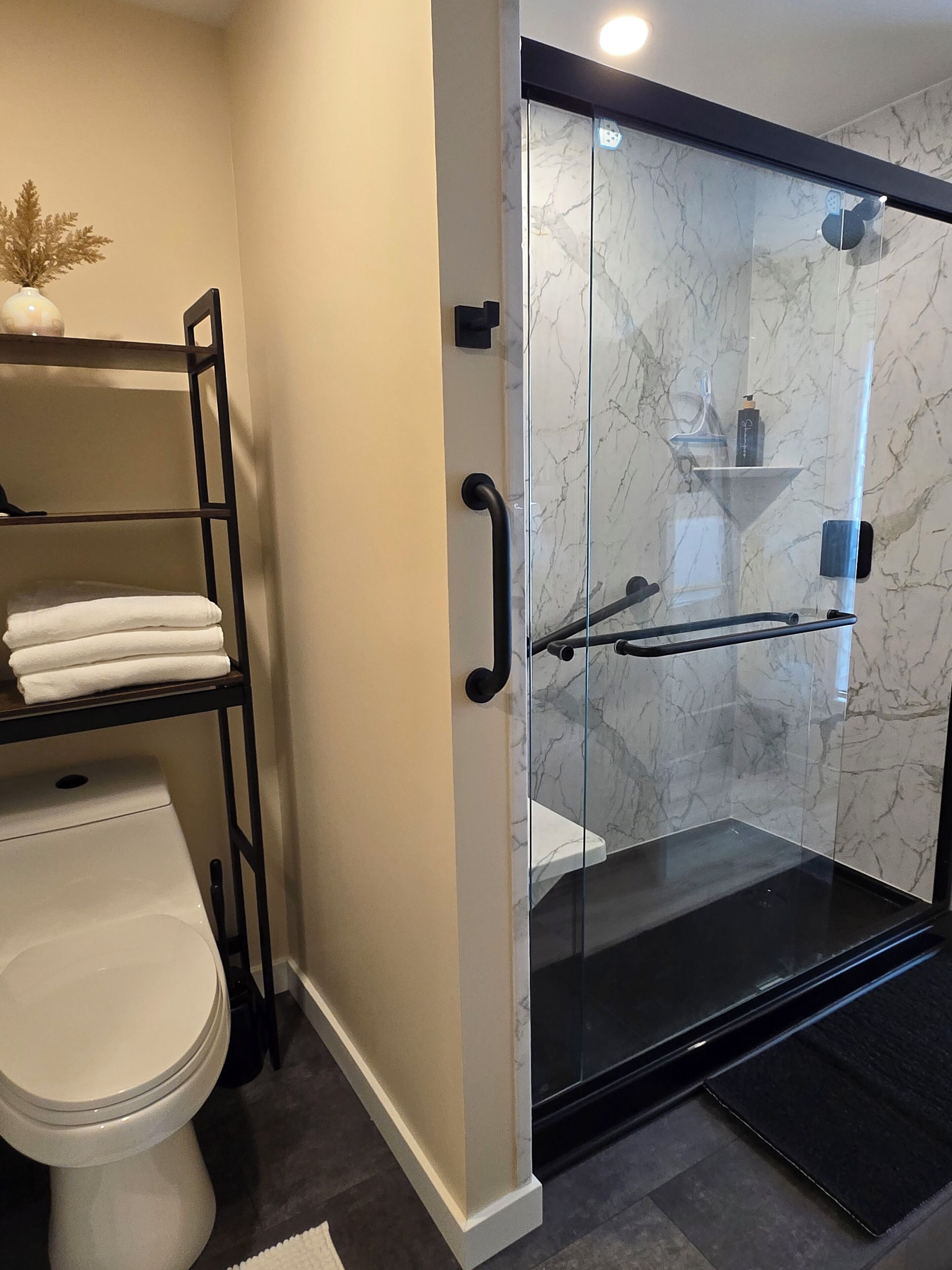 A bathroom with a glass shower enclosure, a toilet, and a shelf with folded towels.