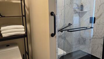 A bathroom with a glass shower enclosure, a toilet, and a shelf with folded towels.
