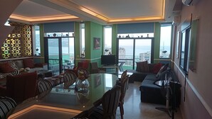 Ein geräumiges Wohnzimmer mit einem Glastisch, bequemen Sitzgelegenheiten, einem großen Fenster mit Meerblick und einem Balkon mit Blick auf die Stadt.