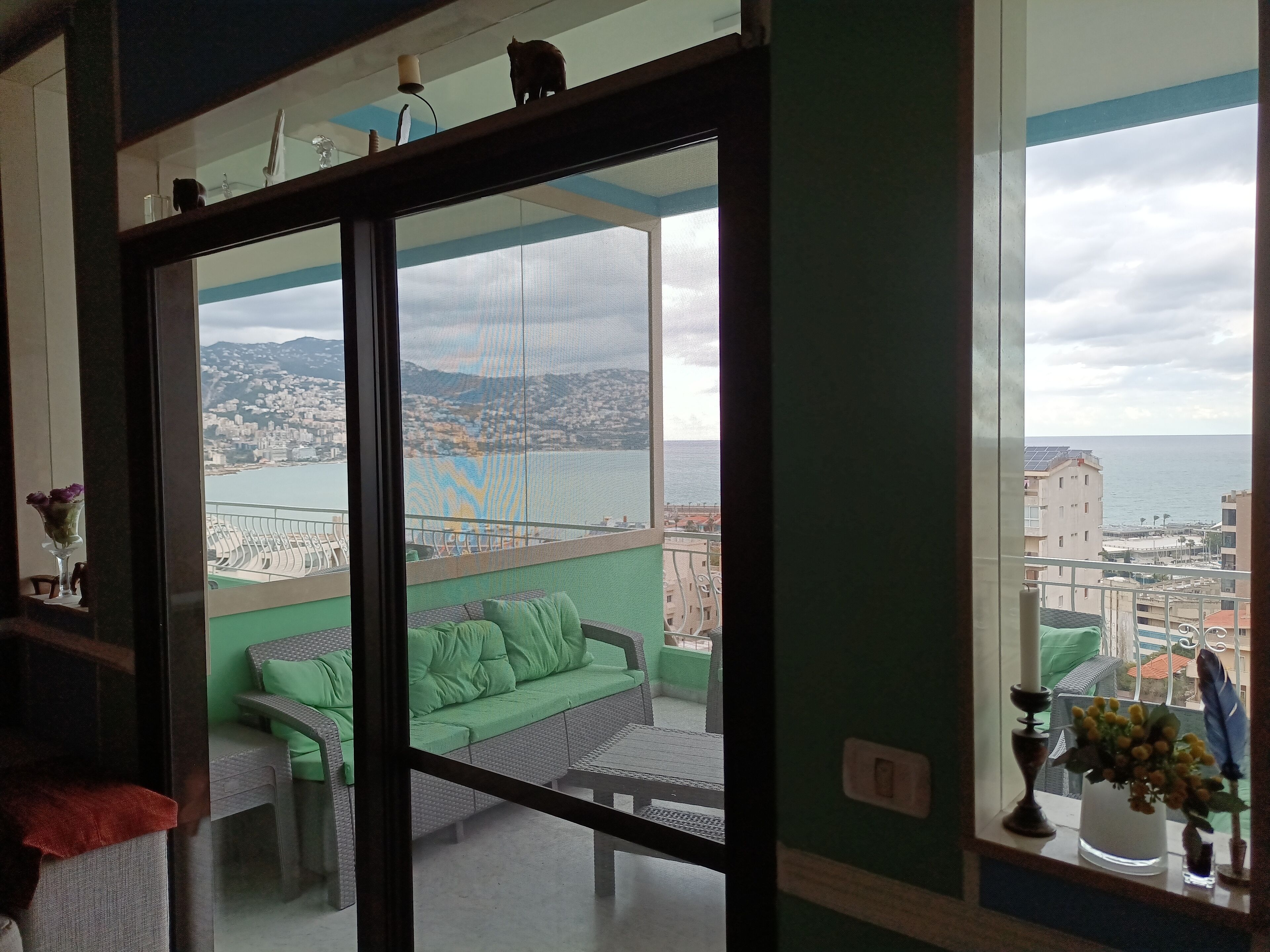 A balcony with a green sofa and a dining table, overlooking a cityscape and the sea.