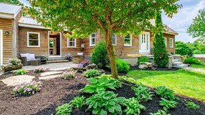 Una casa de madera con un jardín bien cuidado, que incluye un árbol grande y diversas plantas.