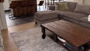 Una sala de estar moderna con un sofá seccional, una mesa de centro con ruedas, una alfombra y una zona de cocina con barra y asientos.