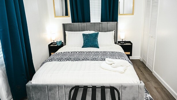 Ein ordentlich bezogenes Bett mit grauem Kopfteil, einer Bettdecke mit weiß-schwarzem Muster und einem türkisfarbenen Kissen. Es gibt zwei Nachttische mit Lampen, einen Spiegel an der Wand und einen weißen Kleiderschrank mit Klappläden.