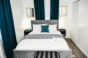 Un lit bien fait, avec une tête de lit grise, une couvre-lit à motifs blancs et noirs, et un oreiller turquoise. On y trouve deux tables de chevet avec des lampes, un miroir au mur et une armoire blanche à portes à persiennes.
