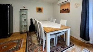 Een eethoek met een houten tafel en witte stoelen, een glazen kast met beeldjes, een kamerkleed met patroon en een klok aan de muur.