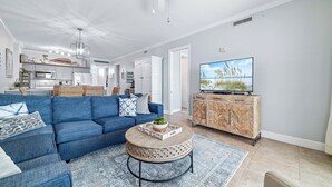 A modern living room with a blue sofa, a round coffee table, and a television on a wooden cabinet. - Grand Pointe 306 | Beachfront, Pools, Saunas, Hot Tub | My Beach Getaways (Orange Beach)