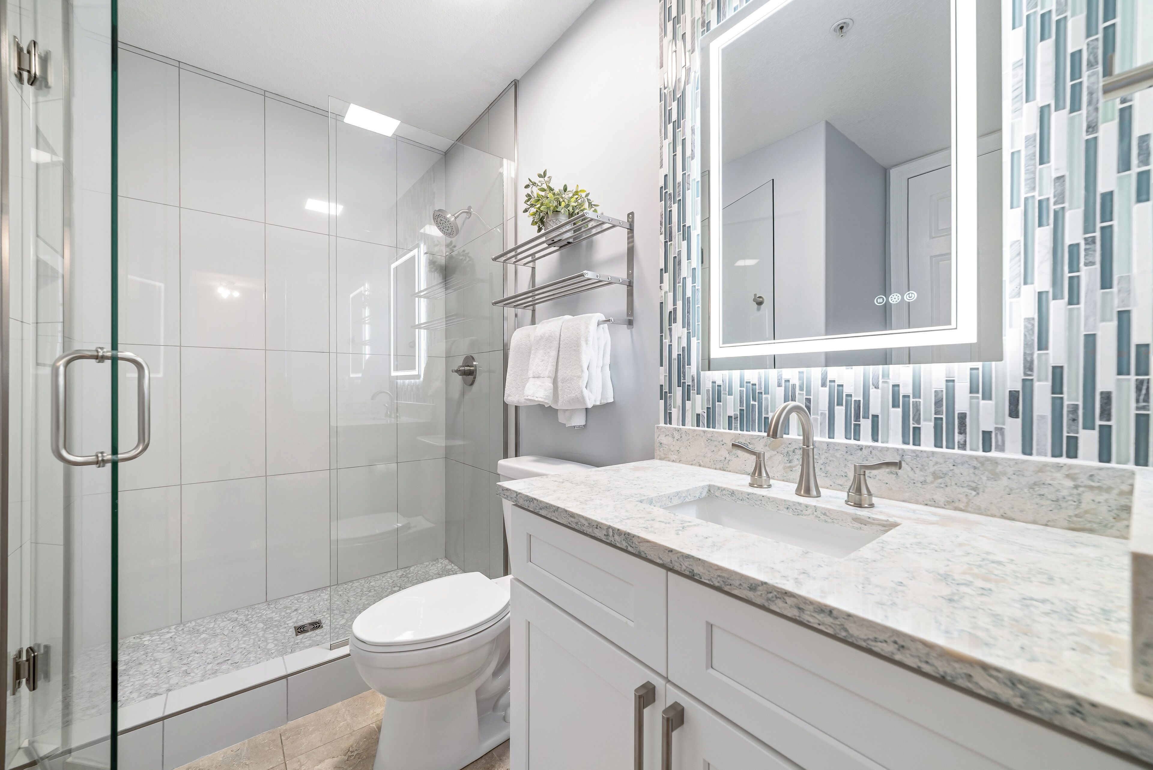 Un baño moderno con cabina de ducha de vidrio, inodoro blanco, encimera de mármol y un espejo grande con resalte de mosaico.