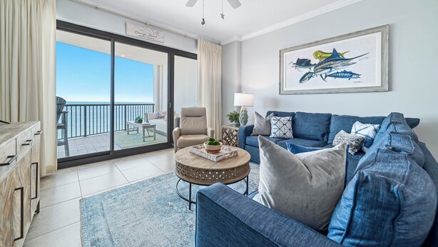 A spacious living room with a large sofa, a coffee table, and a ceiling fan. There is a large window with a view of the ocean. - Grand Pointe 306 | Beachfront, Pools, Saunas, Hot Tub | My Beach Getaways (Orange Beach)