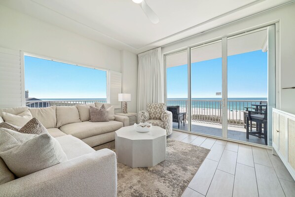 A spacious living room with a large sofa, a round coffee table, and a view of the beach through large sliding doors.