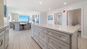 A modern kitchen with a central island, a dining area with a sofa, and a large window offering a view. - Spanish Key 301 | Steps from Flora Bama | Pools, Hot Tub | My Beach Getaways (Pensacola)