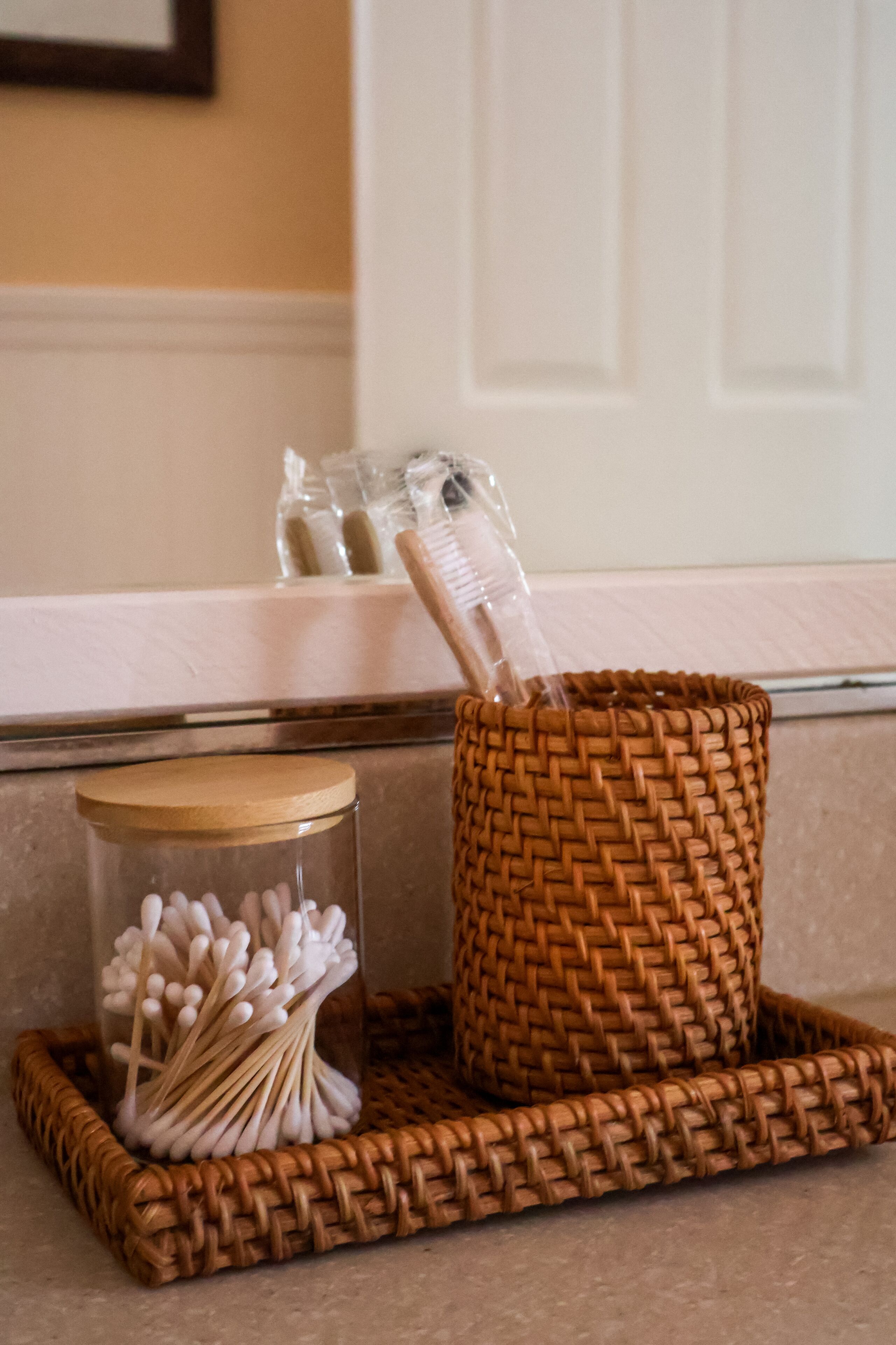 A woven basket on a tray with cotton swabs and a glass jar with a wooden lid.