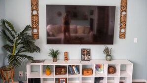 A living room with a television mounted on the wall, a white entertainment unit below, and a large potted plant to the left.