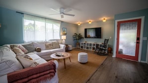 A living room with a white sofa, a coffee table, a rug, a ceiling fan, a TV, and a red-framed door.