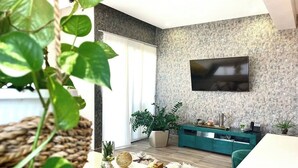 A modern living room with a flat-screen TV mounted on a textured wall, a teal console table, a white sofa, and a dining area with a white table and green chairs. - NewExclusive Apartment Downtown, GYM, POOL&MORE 407 (Santo Domingo)