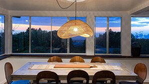A modern dining room with a large table and chairs, a hanging light fixture, and large windows offering a view of the outdoors. - Lihau Boho House (Kailua-Kona)