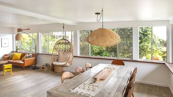 A spacious living area with a large wooden dining table, comfortable seating, and large windows offering a view of greenery.