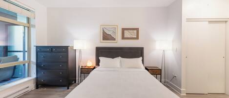 A bedroom with a large bed, a dresser, two bedside lamps, and a window with blinds.