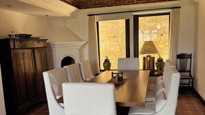 Een eethoek met een houten tafel, witte fauteuils, een bakstenen plafond en uitzicht door een raam.