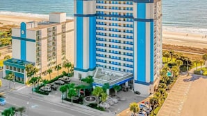 A hotel with a central tower, a parking lot, and a beach view.