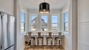 Une cuisine équipée d’un réfrigérateur en acier inoxydable, de quatre chaises en métal, d’un luminaire suspendu et de grandes fenêtres donnant sur des maisons.