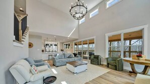 Un vaste salon comprenant un grand canapé, une table basse et un coin repas avec une table en bois. Un escalier avec une rampe se trouve à gauche, et de grandes fenêtres à droite.