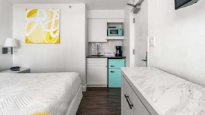 Habitació d'hotel senzilla amb llit, una cuineta petita, televisió penjada a la paret i una taula de nit amb làmpada.