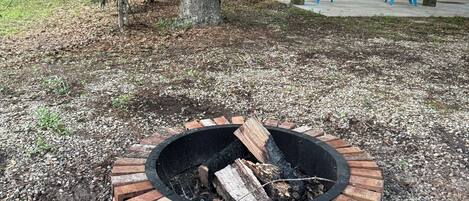 A circular brick fire pit with logs, surrounded by gravel, in a backyard with a wooden deck and blue chairs.