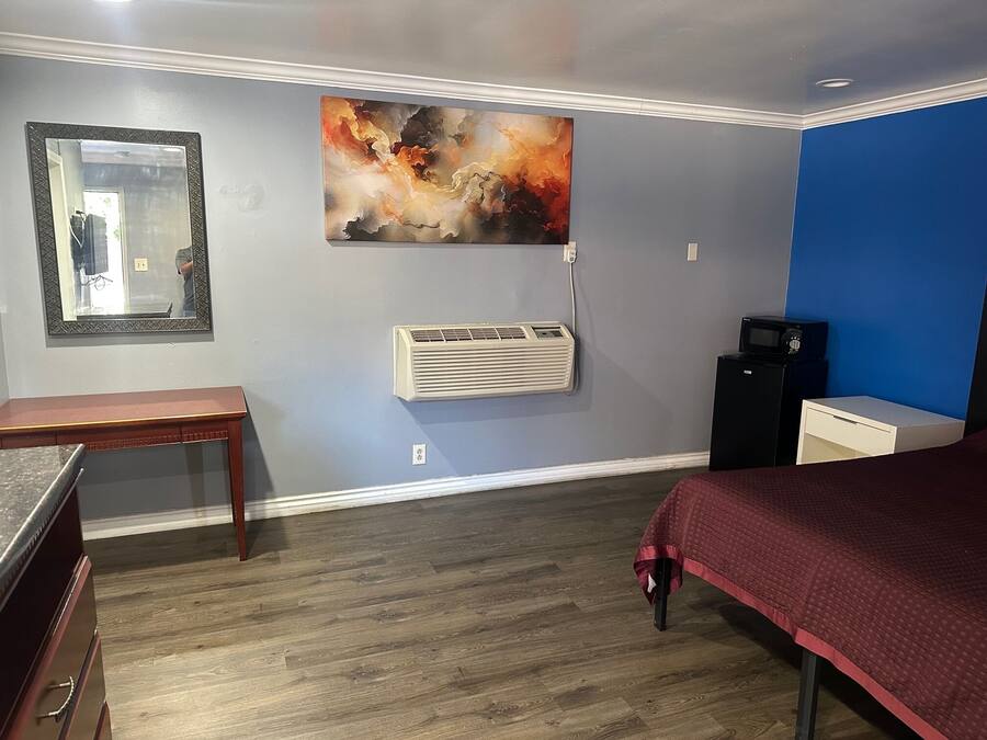 Een hotelkamer met een houten vloer, een bed met een rode deken, een bureau, een stoel, een groot abstract schilderij, een airconditioningapparaat en een zwarte speaker.