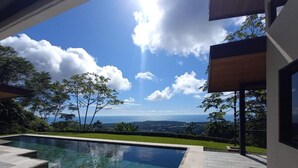 A modern outdoor pool area with clear blue water, surrounded by a stone patio, and offering a scenic view of the ocean and hills.