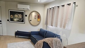 A modern living room with a blue sofa, a round mirror, a white air conditioner, and a window with beige curtains. - Spacious 2BR modern living in Central Eastside location (Los Angeles)