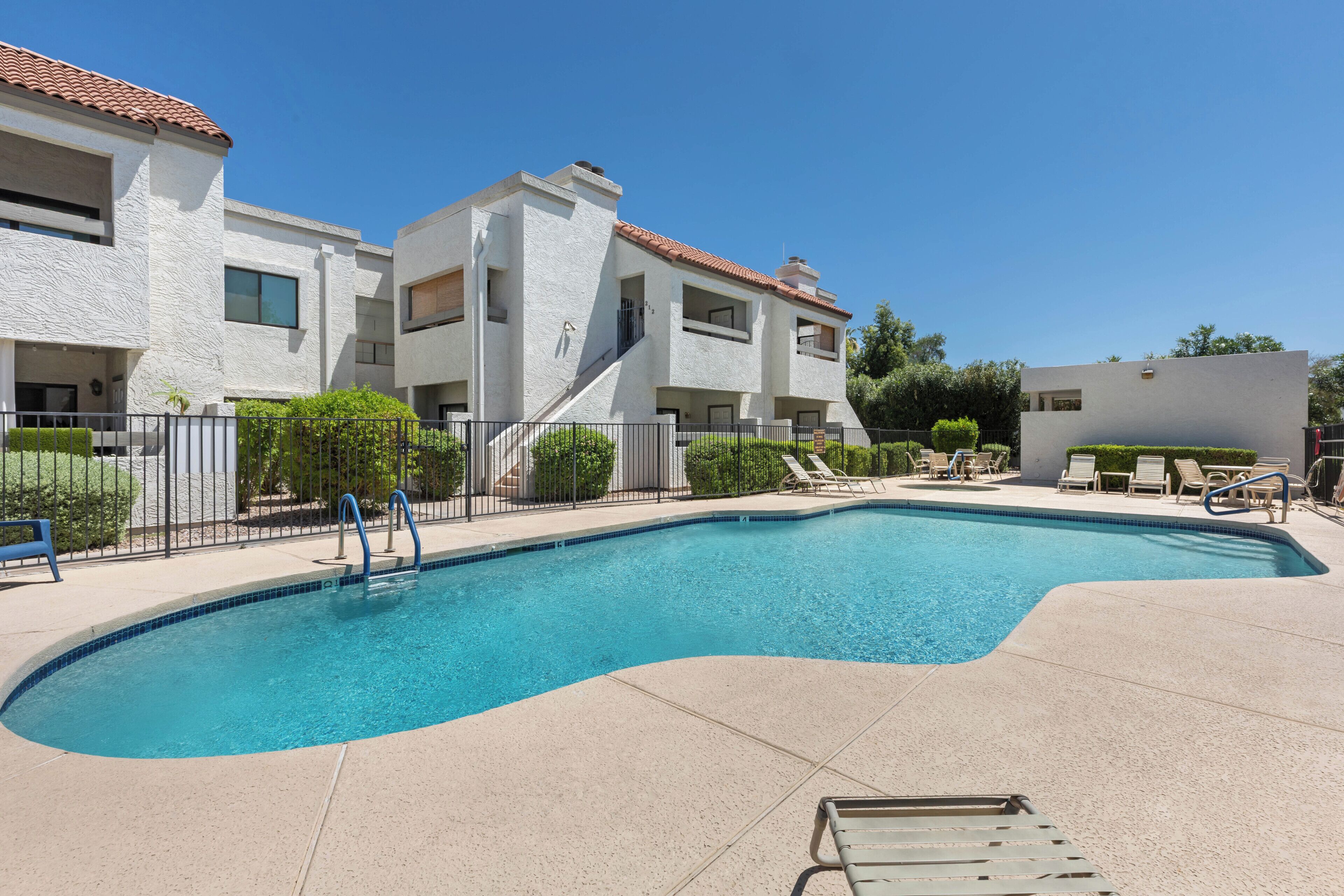A swimming pool with a curved edge, surrounded by a paved deck. There are lounge chairs and a small staircase leading to a building with a tiled roof.