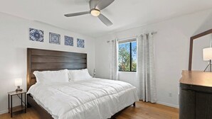 A bedroom with a wooden bed, a ceiling fan, a nightstand with a lamp, and a window with curtains. - Modern Scottsdale Condo with Private Patio, Community Pool and Hot Tub (Scottsdale)