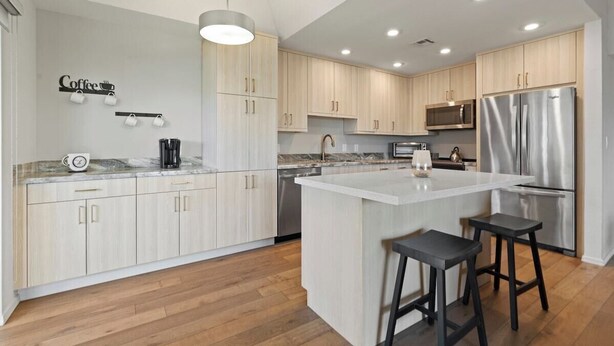 A modern kitchen with light wood cabinets, a large island with stools, stainless steel appliances, and a coffee-themed wall decoration. - Modern Scottsdale Condo with Private Patio, Community Pool and Hot Tub (Scottsdale)