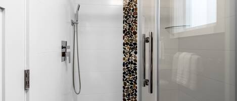 En brusekabine med glasvægge, væg af småsten i mosaik, en glasshylde og et håndklædetørrestativ.