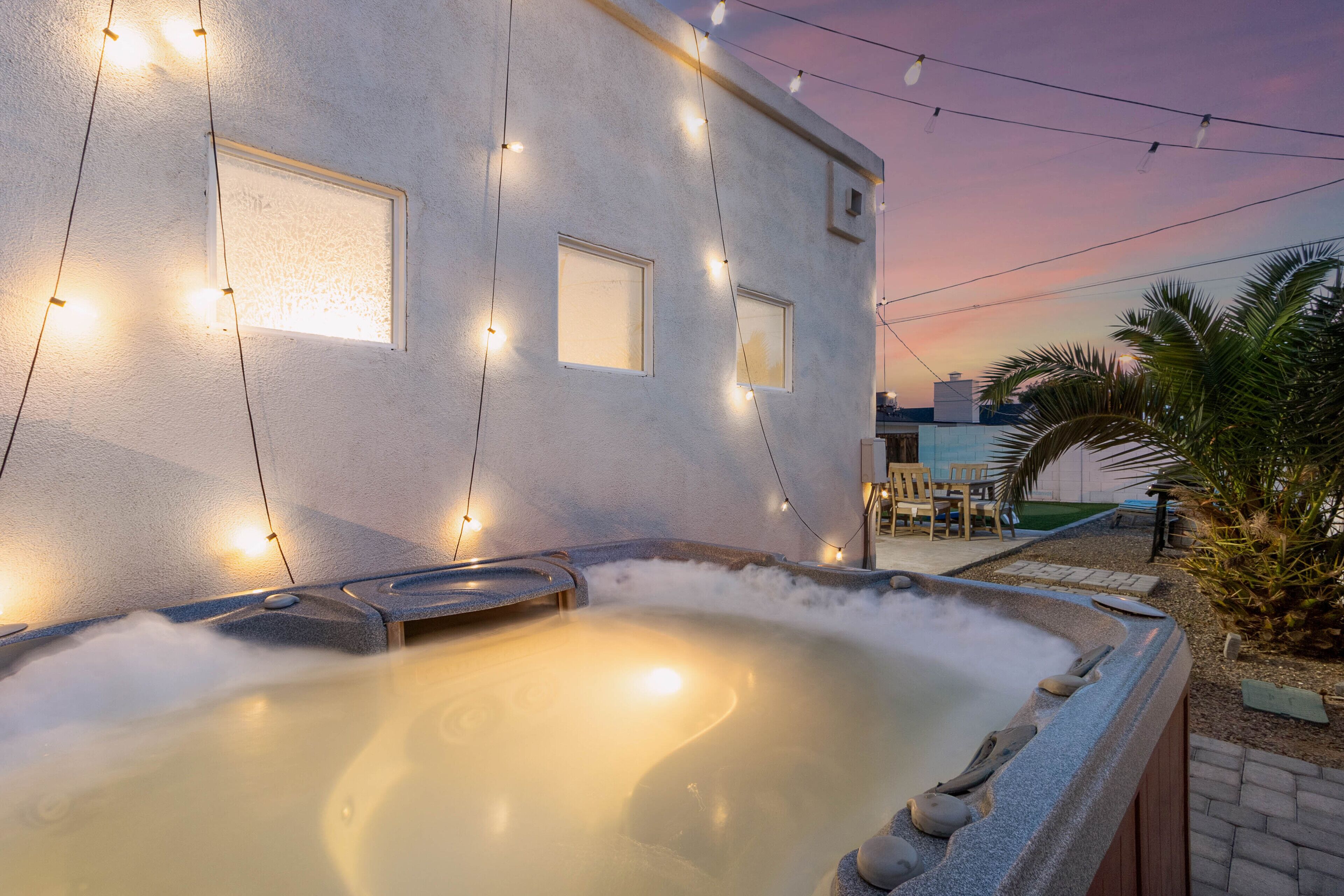 A hot tub with string lights against a building, surrounded by palm trees and a patio area.