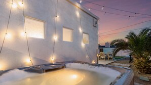 Un jacuzzi iluminado con luces colgantes frente a un edificio, rodeado de palmeras y un área de patio.
